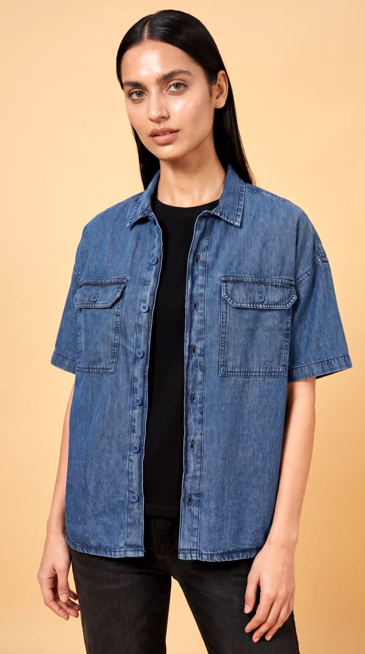 Short sleeve denim shirt with two flap pockets, worn open over a black tee by a woman AI model on a peach backdrop